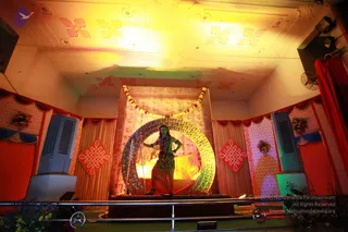 A decorated stage set inside a hall with a patterned ceiling lit in warm yellow light. A standing performer in a green-faced costume with a red and green skirt and a green garland stands before a large golden circular ring framed by a bright glowing backdrop, with a flower garland arching above. A trident-like staff leans near the figure. Red draped curtains, a red diamond-shaped motif and floral arrangements decorate the sides, and two large cooling units stand on either side. A metal railing runs along the front of the stage.