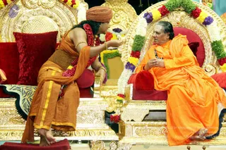 Two people sit on ornate gold-carved thrones with red velvet cushions and garlands of white, yellow, purple and red flowers. On the left, a woman in orange sari-style clothing with a gold waist band, a red-and-orange head wrap, red flower garland and gold bangles leans forward and gestures toward the seated figure on the right. On the right, an elder with grey hair and a rudraksha mala wears a flowing saffron-orange robe and sits with hands together in front of him. Swamiji (probable): the seated elder has long grey swept-back hair, saffron robes and a rudraksha mala, in keeping with the page's description of a satsang. A small red cushion rests on the gold platform below.