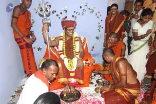 A seated figure in a red-brown robe with an orange-and-gold border and a red-orange turban sits at the centre, smiling, holding a silver trident and wearing a long white and yellow-green garland with a red flower garland below. A candidate for a person is shown with distinctive ornate cues including a crown-like turban, ceremonial gold-trimmed robes, rudraksha beads and garlands. Their feet rest on a brass tray of fruit and flowers set on pink petals, while a shaved-headed man in an orange-gold dhoti and rudraksha mala kneels at right pouring from a brass vessel, and a man in a white shirt and red shawl crouches at lower left. Behind them, a man in an orange dhoti stands at left, a bare-chested young man in orange sits, and women in orange and red saris and a woman in a white sari with green border stand at right. The wall is pale blue with red roses pinned to it, and brass lamps and bowls sit on a white cloth.