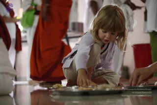 A young blonde-haired girl crouches on a polished floor, reaching toward a steel plate of food, possibly prasadam or annadan offerings. She wears a white long-sleeved top, striped cream trousers, and a dark bead necklace. Behind her, a standing figure wears a flowing orange-and-red garment, and other people in white clothes stand nearby. The background shows a light-coloured room with a red cloth draped on a seat at right.