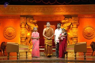A photograph of a stage set with glowing orange and gold carved panels, lion sculptures and a red velvet throne-style seat in the background. Four people stand centrally on the stage. A man in the centre wears a saffron-orange dhoti and shawl with a long beaded necklace and stands with hands folded; he is not clearly identifiable from appearance, so no a person candidate is asserted. To his left an elderly woman wears a pink sari; to his right a man in a white shirt holds a paper, and a woman in a red sari holds a baby wrapped in pink. Two wooden lecterns with red-cushioned seats flank the group.