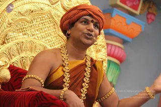 A man seated on an ornate gold throne speaks while gesturing with his right hand. He wears an orange-red turban with a red bindi on his forehead, an orange and yellow shawl draped over bare shoulders, a long gold necklace of ornate beads, and gold bangles on both upper arms. A red and gold striped cushion lies beside him, and colourful orange, pink and green decorative pillars stand behind him. A small lapel microphone is clipped near his mouth. Swamiji (probable): the visible turban, saffron shawl, ceremonial gold necklace and bangles match the page's note that he is present, so he is treated as present on the throne during a discourse.
