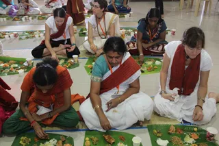 Photograph of a seated communal meal in a tiled hall with devotees sitting cross-legged on mats before banana leaf plates heaped with rice, curries, fried items and pickles, with paper cups of drink nearby. In the foreground a woman in an orange top and green trousers, wearing a red shawl and orange lanyard, eats with her fingers; beside her a woman in a white sari with a red shawl and gold bangles eats from her leaf; at right a woman in a white top with a red shawl and red wrist thread eats while a woman in a teal blouse with a red shawl sits behind. Several other devotees in white and red shawls are seated further back. No a person candidate is visibly present.
