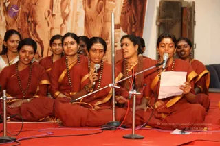 A group of women in maroon and rust-red saris with gold borders sit cross-legged on a red carpet, singing into microphones on stands. Several wear rudraksha malas and forehead markings. The two front singers hold microphones, and a woman at right holds a sheet of paper and reads from it. Behind them a large painted mural of temple carvings and figures covers the wall, with a wooden door at upper right. No a person figure is visible.