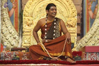 A man with long swept-back dark hair and a red tilak on his forehead sits cross-legged on a red cushioned dais in front of an ornate gold throne-back with carved lion motifs. He wears a rust-orange wrap with a leopard-print shawl draped over one shoulder, and a long mala of rudraksha and white beads hangs at his chest. He holds a rope-like cord in his hands and smiles. Swamiji (probable): the seated man has long swept-back hair, a tilak, rudraksha mala and orange garments.