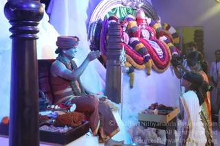 A photograph of a seated man in an ornate ceremonial setting, dressed in a rust-coloured turban and rust-and-gold draped garment with a dark rudraksha-style mala around his neck. He sits on a dark wooden throne-like seat, extending his hand toward a young attendant in white and gold kneeling beside a tray of fruit and offerings. Behind him stands a large deity murti decorated with garlands in red, purple, white and yellow flowers, with a trident placed before it. A photographer with a camera stands at right, and a few onlookers are partly visible at the edges. A dark wooden pillar and white draped backdrop frame the scene, and a silver trident and small brass vessels sit near the seat.