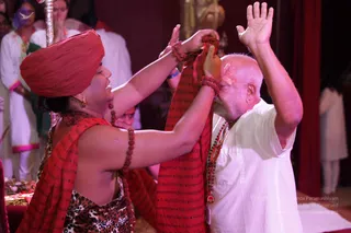 A photograph of a ritual exchange in a warmly lit hall with red-curtained background. A woman in a red turban and red shawl printed with script, wearing a leopard-print blouse, rudraksha beads and gold earrings, holds a red shawl around the head and shoulders of an older man in a white kurta with a rudraksha mala, who bows forward with his hands raised. Two other people in white clothing stand behind them, and offerings and a brass lamp sit at the lower left.