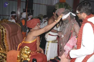 A photograph inside a tent-like hall at a Kumbh Mela camp. A seated figure in an orange-red turban and saffron robe with a gold-edged shawl, seated on an ornate brown leather throne with gold carved armrests, extends his right hand to touch the forehead of a standing sadhu with long grey matted hair, a grey beard, layered rudraksha malas and a white headband. The sadhu wears pink and white cloth. Behind them, attendants in white dhotis and white headbands with gold-banded waistcloths stand near a green and gold canopy. A man in white with glasses and a red-and-white shawl stands at right. Seated devotees in orange and white are visible at left, with a gold lion-footed armrest in the foreground.
