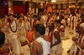A crowded indoor hall with cream marble floors and red walls, where many men in white dhoti-style clothes with gold borders move through the room in a dense group. Many wear red-and-maroon shawls and lanyards with green badges. Several hold small colourful sticks or wands, and one man in the foreground carries a red-and-gold drum. Tall decorative pillars in orange, green and red hang from the ceiling, and a gold tree stands at the back. The scene resembles a procession or group devotional movement. No single person is identifiable as a person here.