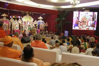 A ceremonial hall with red drapes hosts a puja before an elaborately decorated altar with golden canopies, a green-draped dais, and deity murti in gilded ornaments. Priests in white dhotis and orange-clad sannyasis sit on the floor performing ritual before the altar, with brass vessels and offerings in front. Seated devotees in white and orange sit on white sofas in the foreground, a woman in a green sari among them. A large screen on the right shows a priest in blue-trimmed white cloth attending the altar. A potted banana plant and chandelier complete the scene.