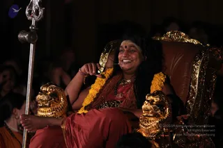 A photograph of a seated person on an ornate golden-and-brown throne with carved golden lion-head armrests, holding a silver trident-topped staff at left. The person wears a red-orange robe, a leopard-print cloth at the waist, rudraksha beads, a gold pendant necklace, a marigold garland, red and white forehead markings and a red bindi, smiling and gesturing with a hand near the mouth. Several audience members are blurred in the dark background. a person seated in a ceremonial chair; the throne, garland and saffron-toned robe with rudraksha are the qualifying cues.