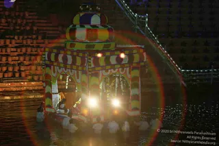 Night photograph of a richly decorated floating shrine barge on a water tank, its tiered, striped canopy of multicoloured blocks topped by a small bell cluster. Floral garlands hang within the open arches and a lit figure sits inside under warm lamplight. Several men in white dhotis and shirts stand waist-deep in the water pushing or guiding the barge. Brick steps lined with potted plants and a lit railing rise behind. No clearly identifiable a person candidate is distinguishable by appearance; the figure seated inside is too small and partly obscured to assess.