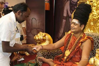 A seated figure in orange-saffron robes with a tall black turban-like headwrap, white forehead marking and red bindi sits on a gold-and-leopard-print throne with a golden lion-head armrest. Layered rudraksha malas and a large gold pendant hang at the chest. He extends a hand toward a man in a white shirt who is handing him a small white packet or card. A camera operator and another man stand behind, and a gold-framed backdrop with a lotus motif is visible.