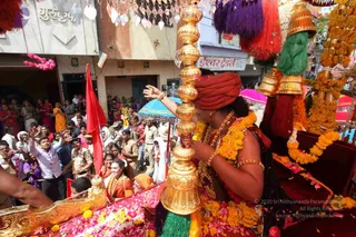 A procession on a city street with a figure in a red-orange turban and orange and gold garments standing on a decorated golden palanquin or float. The figure raises a hand in greeting and wears several long garlands of orange marigolds and a rudraksha-style necklace with bangles. A tall gold ornamental staff with a tapering gold finial stands beside them, and red, purple and green tassels hang from the canopy. Crowds line the street behind, with uniformed police officers in khaki, men in white shirts waving, women in saffron and colourful saris, and red flags. Rose petals and marigold petals cover the float's pink cloth. Shop fronts with signs line the street.