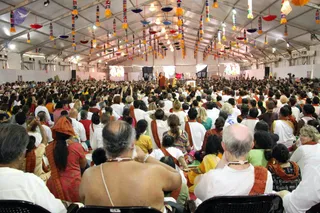 A large photograph taken inside a white tented pavilion hung with colourful tassel decorations and hanging lamps. A dense crowd of several hundred people sits on chairs facing a raised stage at the far end, where a figure in orange robes stands with arms raised, addressing the gathering. In the foreground, several people face away from the camera: a bald man bare-shouldered with a white sacred thread, a man and woman in white clothes with orange-brown shawls, and a woman in a red garment with an orange head wrap. Video screens flank the stage. The crowd is mostly dressed in white with orange shawls.