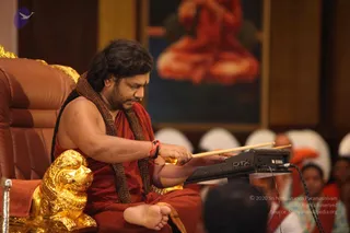 A man with long dark hair, a forehead tilak and a rudraksha mala draped over his shoulders sits on a brown leather throne with gold lion-head armrests. He is bare-chested, wearing a red-orange cloth wrapped around his waist and legs, and plays a drum pad with wooden sticks on a black electronic percussion unit. Behind him a large painting shows a red-robed seated figure in meditation. Audience members in white and orange clothing sit in the background, and a woman in a dark top and a partly visible person in the foreground frame the scene. The setting is a formal satsang hall with wood-panelled walls.