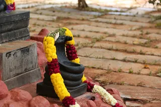 A close view of a black stone shivalinga-style ritual object, more precisely a black stone yoni-pitha base with a curved spout, garlanded with yellow marigolds, red roses and white flowers draped over its top. The garland sweeps down the front and around the base. Beside it stands a grey stone pedestal or plinth, and the ground is a red-brown paved walkway with grass growing between the stones. No people are visible; the scene is a quiet outdoor shrine detail.