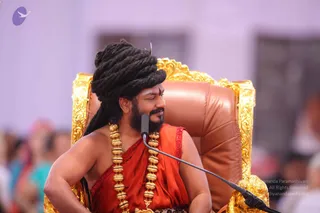 A man seated on an ornate gold-framed brown leather throne, smiling and turning his head to his right toward an off-camera listener. He has a large black dreadlocked crown of hair wrapped high on his head, a short beard, and a red tilak mark on his forehead. He wears an orange-red sleeveless garment with a red-patterned border and heavy gold ceremonial necklaces of bead-like links. A microphone on a black stand is in front of him. The blurred background shows a white wall and out-of-focus audience members in colourful clothing.