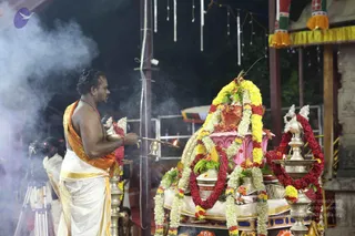 A shaved-headed priest in a white dhoti with an orange-gold shawl and a red rudraksha-style mala stands bare-chested at left, holding a brass lamp or incense vessel from which smoke rises. Before him a decorated ritual arrangement is adorned with yellow and red marigold garlands, green leaves and a pink-covered base, with brass lamps on either side. Smoke drifts across the frame under a dark night sky, with hanging string decorations and yellow-fringed canopy above. Several other figures are partially visible at the edges.