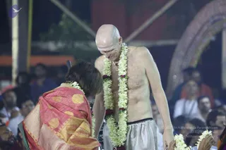A bald, bare-chested man stands at night wearing white draped lower garments and a long double garland of white flowers with red blossoms. A woman in a red and gold patterned sari bends forward toward him, her hands near his. Behind them a crowd watches, and a decorated arch and a red structure are visible.
