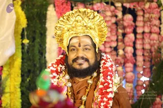 A bearded man in a large ornate gold crown with a radiating floral-patterned headdress sits before a backdrop of garlands of pink and red flowers and yellow marigold strands. He wears a red-and-white rose garland, an ochre-brown shawl, and heavy ceremonial gold-toned beads and necklaces, with a red tilak mark and white vibhuti on his forehead. The Swamiji (probable) cues are the ornate gold crown, ochre-saffron garments and ceremonial beads, shown in a decorated ritual setting.