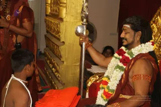 A seated man with long dark hair, a white forehead mark and a garland of white jasmine and red roses sits on a gold throne. He wears a maroon and gold cloth, orange-red armlets and gold earrings, and holds a silver staff topped with a round silver finial. A shirtless young boy in a white sacred thread sits before him, facing him. Behind them, a standing attendant in orange-red clothes holds a microphone, and a gold-carved wooden pillar stands in the background. The setting resembles a ritual or satsang hall.