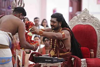 A photograph of a ceremonial moment in a hall with a red-cushioned silver throne on the right. A man with long dreadlocked hair, wearing a maroon-red robe, gold beaded necklaces, gold armbands and a gold bracelet, sits on the throne and reaches for a garland of red and white flowers. Facing him on the left, a bare-chested priest in white dhoti with a blue-edged cloth holds a silver tray with the garland. Behind them stand several onlookers, and a silver trident and a small electronic keyboard sit near the throne. The throned figure's appearance shows several cues (long dreadlocked hair, ornate gold jewellery and maroon-red robes), so he is recorded as a person seated in person.