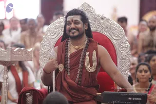 A smiling man with long dark hair and a tilak on his forehead sits on an ornate silver throne with a red cushion, wearing a rust-red saffron wrap, a crystal and rudraksha mala, a gold pendant necklace and a large white beaded garland across his chest. He holds a silver ceremonial staff in one hand. Behind him and around the throne, many seated devotees in white and orange clothing watch, and a black Yamaha keyboard sits in the foreground. Several people are visible in the background audience.