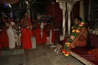 A photograph of a nighttime ceremony in an open courtyard under string lights. On the right, a man with a shaved head and tilak sits smiling on an ornate silver-and-red throne-like seat, wearing a saffron-orange robe and a long garland of green leaves with red and yellow flowers. Several young men in saffron-orange dhotis, some bare-chested and wearing rudraksha malas, stand in a row with hands folded in namaskar, and one holds a red-and-gold cloth tray. A white-clad woman and other white-clad attendants stand at left near a brass oil lamp on a stand. Silver pillars with hanging chains frame the seat, and a red wall rises behind the group.