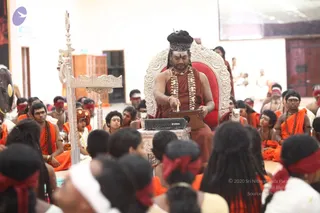 A seated man in a central ceremonial position on an ornate silver-and-red throne, wearing an orange-red shawl over bare shoulders, a layered rudraksha and pearl-type mala, and a tall swept-back hairstyle with a decorated crown-like band. He holds a small wooden tray or board and a stylus-like object, with a Yamaha keyboard unit on a stand before him. Around him sit a large group of young initiates in saffron-orange clothing and rudraksha malas, many wearing red blindfold cloths over their eyes. A tall silver ceremonial lamp stand stands to the left. Walls are white with a wooden door and a large screen at the top right. The image is a photograph of a seated ritual gathering. Swamiji (probable) cues: the central figure has a swept-back hairstyle with a crown, a shawl over bare shoulders and ornate mala necklaces, at a ceremonial throne.