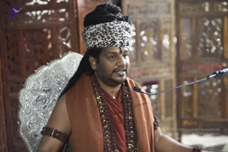 A photograph of a man seated before a microphone on a stand, speaking, in an ornately carved wooden-screened interior. He wears a leopard-print cloth wrapped as a turban over long dark hair, with a dark tilak on his forehead, a rust-orange shawl over a matching orange garment, and multiple strands of brown rudraksha beads with gold-coloured spacers around his neck. A silver-embossed ornamental seat stands behind him, and a rudraksha bracelet is on his left forearm. Swamiji (probable) appears as the speaker, with the cues of long hair under a turban, orange clothing and rudraksha malas. The page is about an Aushada preparation event on 29 August 2017.
