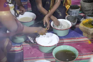 A photograph of a ritual preparation scene on a colourful striped cloth. Several enamel basins with red rims hold white powder, a pale grainy mixture, dark green liquid and yellow turmeric-like powder. Hands in gold-bordered dark clothing mix and press the substances, while a stone mortar with yellow powder and a grinding stone sits at the right edge. A partially visible person in red and gold sits at the left. Other people are visible only as hands and arms.