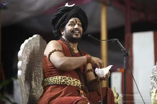 A man seated on an ornate silver throne with a carved backrest, smiling and holding a white conch-shaped vessel in his right hand. He wears a rust-orange robe, a leopard-print waist sash, multiple rudraksha mala strands, rudraksha bracelets and a gold chain. His long black hair is piled into a turban topped with a white crescent ornament, and he has a white tripundra on his forehead. A microphone on a stand is beside him, with a white and red striped curtain and a silver deity figure at the right edge. a person appears in person, seated and speaking to an audience, with the ceremonial clothing and ornaments matching the page's event context.