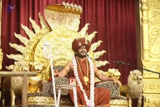A seated man in an orange-saffron turban and matching saffron robes sits on an ornate golden throne carved with a large cobra hood and radiating sun rays, flanked by golden lion figures and leopard-print cushions. He wears a pink and white flower garland, a gold pendant necklace, bangles on both wrists, and holds a white staff in his left hand while gesturing with his right. A microphone stands at his side and a small lectern is in front of him against a deep red curtain. Gilded carved pillars frame the right edge.