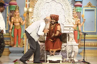 A seated man in an orange-red saffron robe and a matching orange-red turban sits on an ornate silver throne with leopard-print upholstery, smiling and holding his hands together. Rudraksha malas hang around his neck and wrists, and a gold necklace is visible. Swamiji (probable): the seated figure wears saffron clothing, a turban and rudraksha malas, matching the page's claim. A bald, white-bearded elderly man in a grey vest over a white shirt and dark trousers bends forward to kiss or greet him on the cheek. Behind them stand two painted deity murti figures with multiple arms, holding ritual objects, in green and red garments, beside a microphone stand on a decorated stage with a red carpet.