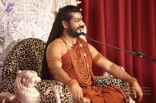 A man with long dark dreadlocks and a short beard sits on an ornate leopard-print throne with silver carved lion-head armrests, in front of a red curtain and a silver embossed backdrop. He wears a rust-orange wrap, multiple strands of rudraksha-style beads across the chest, a gold necklace, and beaded bands on his upper arms and wrists, and speaks into a microphone on a stand. a person: long swept-back hair, ornate gold necklace and rudraksha malas on a saffron-orange robe, seated on a throne. One person is visible.
