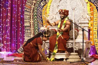 A seated man on a silver ornate throne wearing a rust-orange turban with gold ornament, rust-orange dhoti and shawl, yellow and white flower garlands, and beaded bracelets, holding a gold-tipped staff or rod. A woman in a rust-orange sari kneels on a red-patterned rug at his feet, holding a brass object near the floor. Behind them hang colourful embroidered panels, purple fairy lights and a tall silver stand. A ceremonial padapuja-style honouring is shown. The throne carries leopard-print cushions, and a small offering tray sits to the left.