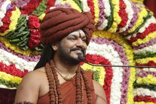 A photograph of a smiling man seated before a microphone stand, in front of a large backdrop of red roses, white and yellow flowers and green leaves. He wears a rust-red turban, a rudraksha mala of dark beads draped over his shoulders, a gold pendant necklace, and a rust-orange wrap over bare shoulders, with a tilak mark on his forehead.