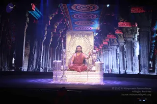A seated figure in orange-red robes sits cross-legged on an ornate golden throne on a stage, holding a long staff in one hand and a rudraksha mala with red beads around the neck. The throne has carved gold lion figures and a gilded arched backrest. Behind him a large projected backdrop shows a stone temple hall with rows of carved pillars and a colourful concentric-patterned ceiling, lit in blue and purple. The stage floor is dark with soft purple light spots. a person: the figure wears a rudraksha mala and orange-saffron robes on a throne, with a long staff, though his face and hair are not clearly visible, so identification is uncertain. Only a small partially visible figure appears in the lower foreground.