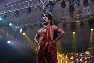 A photograph taken from a low angle on a stage lit by overhead stage lights and yellow spotlights, with metal truss rigging above. A single standing man wears a rust-red dhoti-style garment, a leopard-print shawl draped over one shoulder, rudraksha bracelets on his upper arms and wrists, and a gold ornament on his forehead. His long dark hair is swept back, and he holds his hands on his hips while gazing into the distance. a person cues are present in his long swept-back hair, ornate red-and-gold clothing, shawl and rudraksha adornments, but the appearance is not conclusive enough to confirm identity. A small purple bird logo sits in the upper left corner.