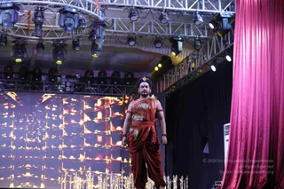 A man stands on a stage in a rust-red and orange draped garment with a leopard-print patterned wrap across the chest, matching red-orange dhoti-style drape, beaded bangles on the arms and a jewelled forehead ornament with dark hair swept back. Behind him a large LED screen shows glowing orange-gold lamp imagery, overhead truss rigging holds stage lights, and a magenta velvet curtain hangs at right. Silver ritual lamps or vessels are set along the front of the stage. Appearance-based identification is not established from the visible cues alone, so the person is described by role as a performer on stage.