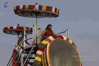 A man in a rust-red sleeveless garment with long dark hair and a tilak on his forehead sits on a decorated float, a large grey stone-like platform garlanded with bands of marigolds, yellow and white flowers and green leaves. Behind him stands a white carved seat. Two multicoloured fringed parasols with gold finials shade the float, and a steel staircase with railings leads up to it. A man in white ritual dress with orange cloth stands on the stairs below. The sky is pale grey.