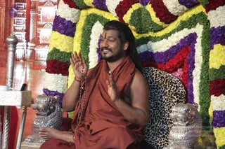 A photograph of a bearded man with long dark hair seated on a leopard-print cushioned throne, barefoot, bare-chested beneath a rust-orange saffron shawl and wearing a rudraksha mala and a forehead tilak. He is smiling and raising both palms in a greeting gesture. Behind him is a backdrop of tiered garlands of red roses, purple and white flowers and green leaves, and silver lion-shaped figures flank the throne. Swamiji (probable): long swept-back hair, saffron shawl, rudraksha mala and ceremonial adornment, seated as the central speaker during a satsang.