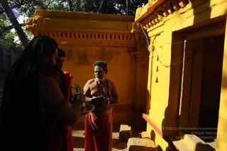 A photograph of an outdoor temple scene beside a yellow-ochre shrine wall with a carved cornice and a small doorway. A bare-chested, grey-bearded man in a red-orange dhoti and a beaded mala stands in the sunlight facing the shrine. To the left, a long-haired man with dreadlocks and another man in orange cloth hold a brass plate of flowers and leaves. Palm fronds overhang the wall.