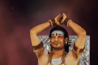 A bare-chested man with swept-back long dark hair sits before an ornate silver throne against a reddish-purple smoky backdrop. He raises both arms overhead with hands joined above his head, in a meditative or ritual pose. He wears rudraksha mala bracelets on his wrists and upper arms, layered gold necklaces with tassels, gold earrings, and white ash markings on his forehead and chest with a dark central bindu. Swamiji (probable) cues: long swept-back hair, rudraksha malas, ceremonial gold necklaces, and distinctive adornments. A faint white bird appears in the upper left.