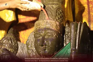 Close photo of a dark stone deity murti being bathed during an abhishekam. A hand at the upper left pours liquid from a white vessel, and a reddish stream runs over the carved crown onto the face of the murti. Carved ornamental patterns cover the headdress and shoulder, and the background shows warm yellow and orange drapery. No people are clearly visible beyond the pouring hand, and no a person candidate appears in the frame.