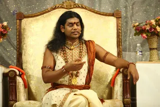 A man with long, swept-back black hair sits on an ornate gold-brocade throne with carved wooden arms and an orange cushion, speaking with one hand raised in a gesture. He wears a cream embroidered kurta with floral patterns, an orange shawl draped over one shoulder, layered rudraksha malas, a gold ceremonial pendant and beaded bracelets, with a red bindi on his forehead. A microphone is clipped near his face. Behind him are a shimmering curtain and pink and white flower arrangements, with a water bottle on a side table. Swamiji (probable): the seated speaker wears long hair, orange shawl, rudraksha malas and ornate gold pendant, consistent with the page's identification of Nithyananda Paramashivam at this event.