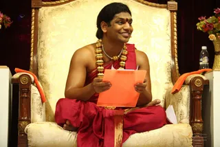 A smiling man with long dark hair and a small red bindi sits cross-legged on an ornate gold and cream upholstered throne with carved wooden armrests. He wears a red-maroon dhoti and a bare-shouldered red shawl, with heavy gold beaded necklaces and a thin silver chain. He holds an orange clipboard with loose papers, and orange cloths drape the armrests. Flower arrangements and a water bottle stand beside the throne.