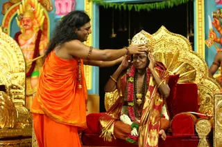 A seated pontiff in a gold crown and red-and-gold silk shawl sits on a red-cushioned golden throne, wearing a rudraksha-style necklace of red flowers and a gold pendant, with white and red garlands draped over his lap. Standing before him, a man with long, swept-back dark hair and a saffron-orange robe places his right hand on the crowned figure's head in a blessing gesture while smiling. Gilded throne backrests and a painted deity figure with a red garland appear behind them, with a green festoon above a blue-and-pink shrine backdrop. The scene reads as an investiture or coronation ceremony in a temple-style hall. Swamiji (probable): the standing man in saffron with long swept-back hair, a rudraksha mala and gold-chain pendant is the.