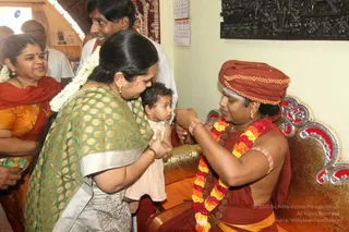 A photograph of a seated man in an orange-red turban and red-orange shawl, bare-chested, wearing a multicolour marigold garland and rudraksha mala, with sacred ash on his forehead, gold-coloured ornate chair with red and silver accents. He holds a thread-like object and reaches toward a small child in a cream dress held by a woman in a green and gold brocade sari with silver border, who leans in. A smiling woman in orange sari with red shawl stands at left, and a man in white shirt stands behind. A carved wooden panel hangs on the wall and a calendar is visible. Figures are in a domestic room setting during a house visit.